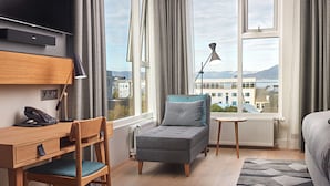 A modern hotel room with a bed, a desk with a chair, a small armchair, a standing lamp, and a view of buildings and mountains through large windows. - Canopy by Hilton Reykjavik City Centre (Reykjavik)