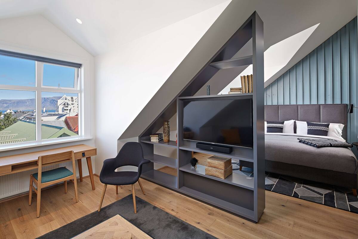 Een moderne loft slaapkamer met een groot raam, een ingebouwd bureau, een comfortabele fauteuil, een bed met hoofdeinde en een televisie op een plank.