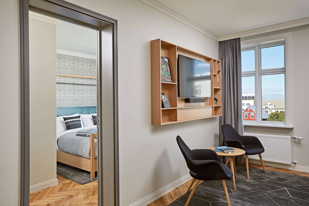 Een moderne kamer met een bed, een aan de muur bevestigde televisie, een klein tafeltje met stoelen en een raam met uitzicht op gebouwen.