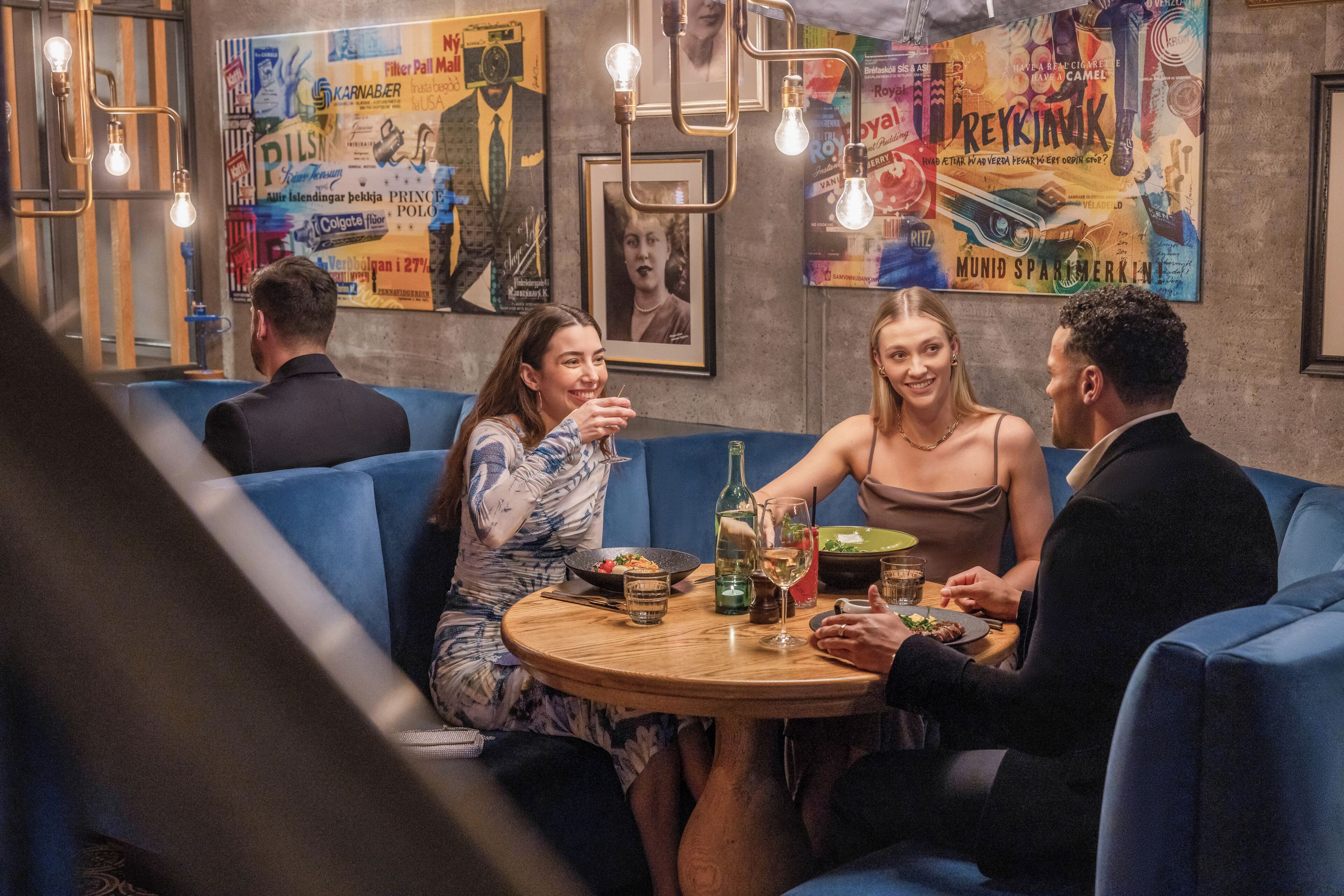 Un groupe de personnes est assis autour d’une table ronde dans un restaurant. Les banquettes sont recouvertes d’un tissu bleu et un mur est décoré de divers affiches.