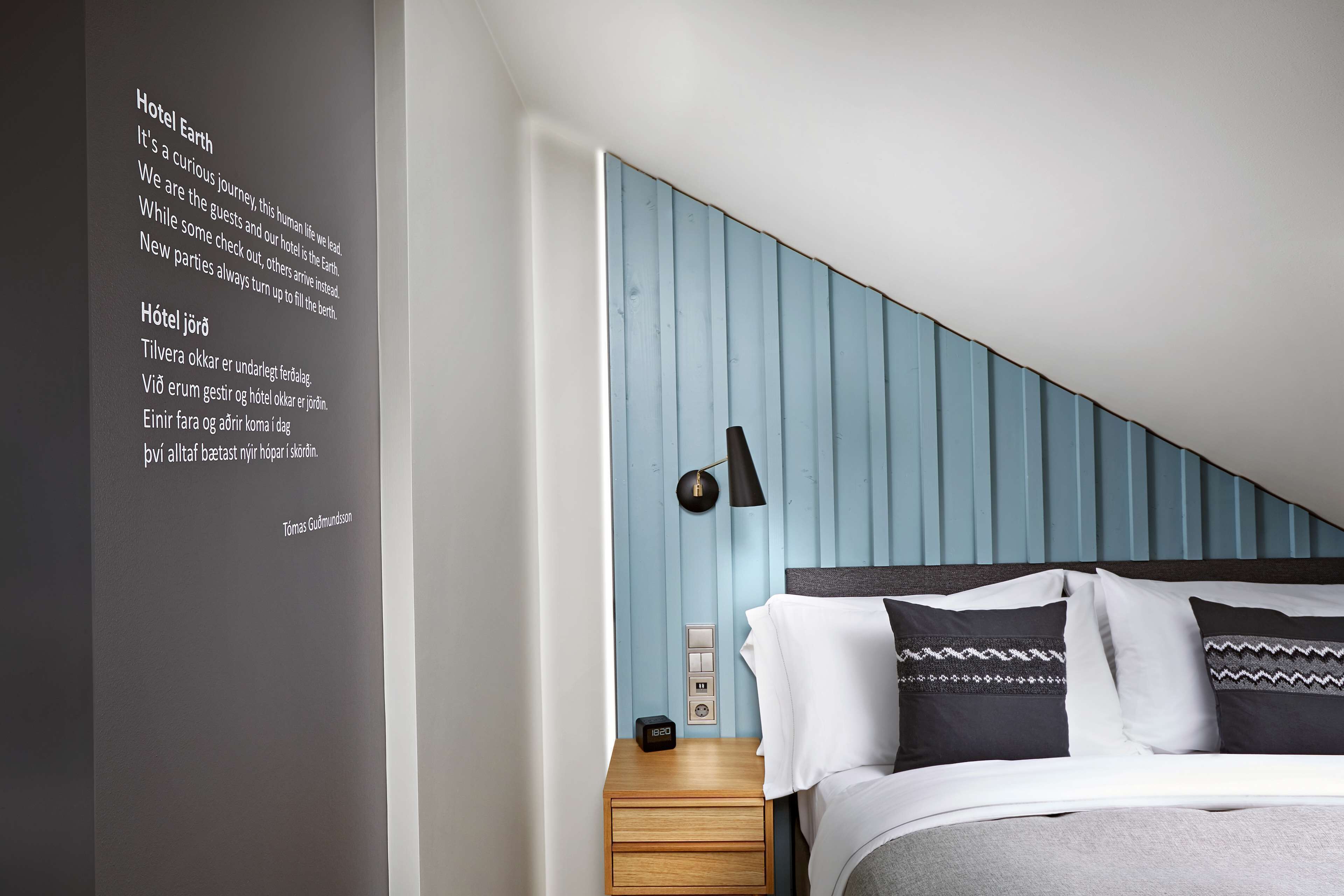 Una habitación de hotel moderna con una pared azul de acento, una cama bien hecha con almohadas estampadas, una mesita de noche con lámpara e interruptor de luz en la pared.