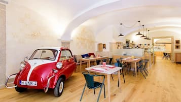 Zona de menjador amb un cotxe antic, taules de fusta, cadires i una cuina de fons.