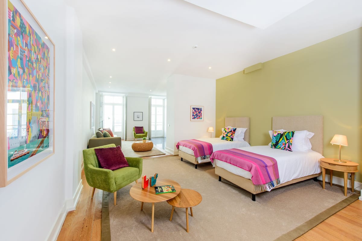 A modern hotel room with a large bed, a green armchair, a small round table, and a colorful artwork on the wall.