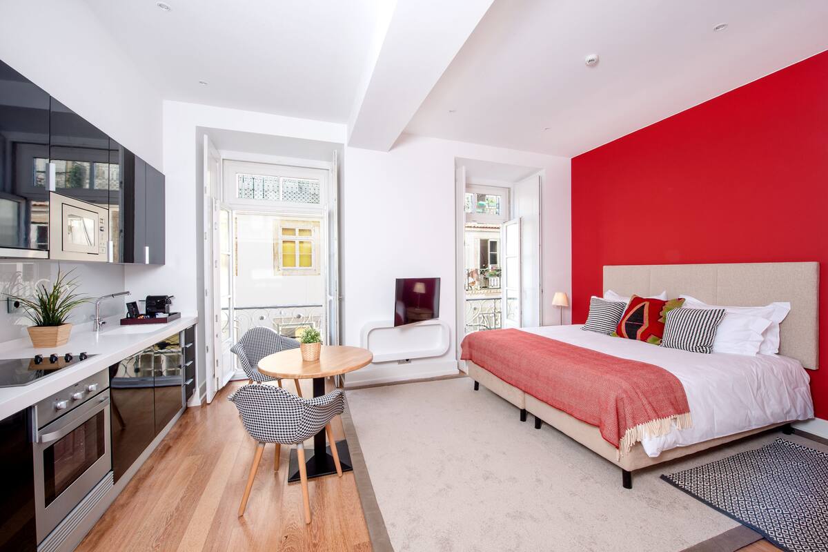 A compact room with a kitchenette, a bed with a red accent wall, and a small seating area.