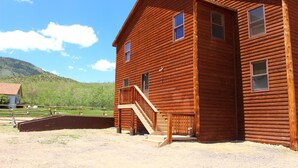 Una cabaña de madera de dos pisos con una escalera que conduce a un balcón, rodeada por una zona vallada con suelo de tierra.