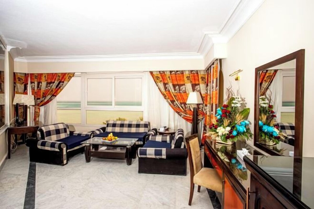 A living room with a sofa set, a coffee table, and a side table. There is a window with curtains, a mirror, and a vase with flowers.