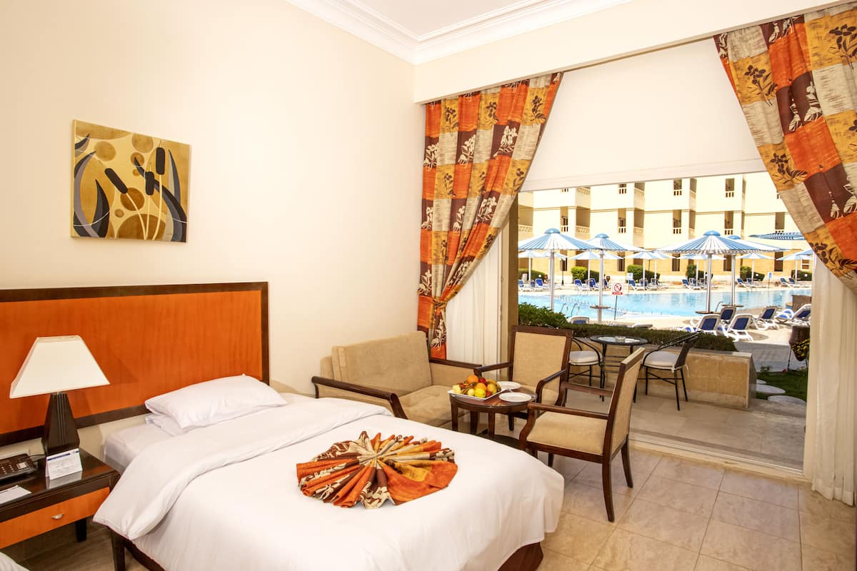 Quarto de hotel com cama, escrivaninha com luminária, cadeira, mesinha com frutas e vista para a piscina e guarda-sóis.