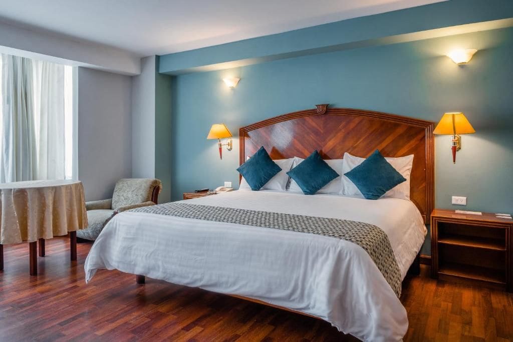 A hotel room with a large bed, two bedside tables, a chair, a small table, and a window with curtains.