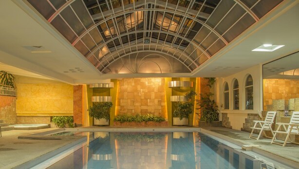 A large indoor swimming pool with a glass ceiling, surrounded by potted plants and seating areas. - Hotel Libertador (Loja)
