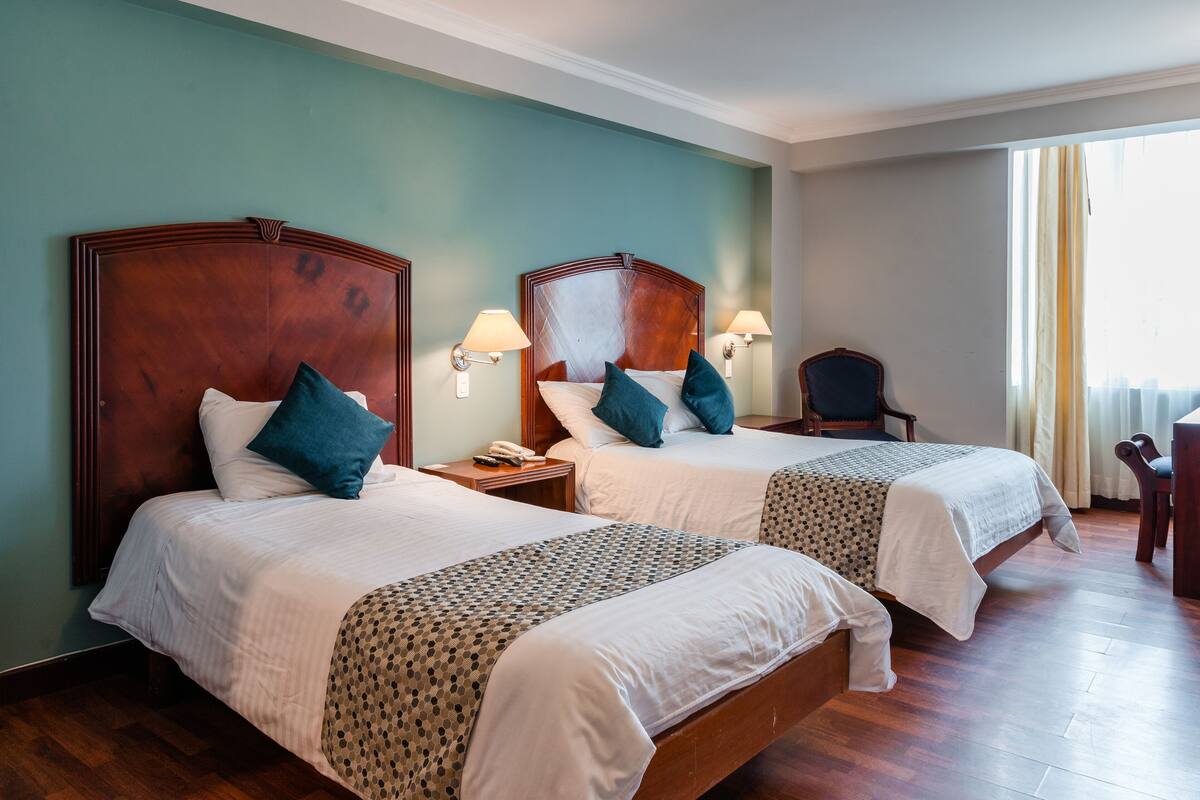 A hotel room with two beds, each with a wooden headboard, white bedding, and teal pillows. There are bedside lamps, a chair, and a desk with a chair.