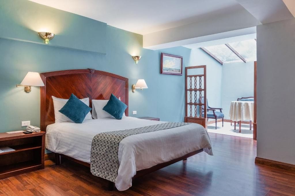 Habitación de hotel con una cama grande, dos lámparas de noche, una mesita de noche, un cabecero de madera, un ventanal con vistas y una pequeña mesa con una silla.