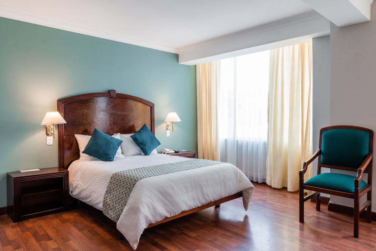 A hotel room with a large bed, wooden headboard, two bedside lamps, a chair, and a window with curtains.