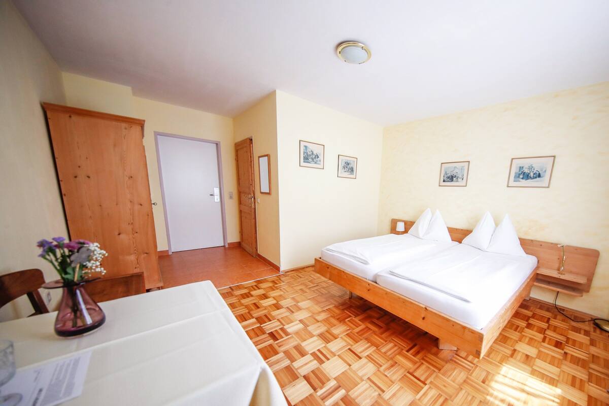 A room with a wooden floor, a bed with white bedding, a wooden door, and a small table with a vase of flowers.