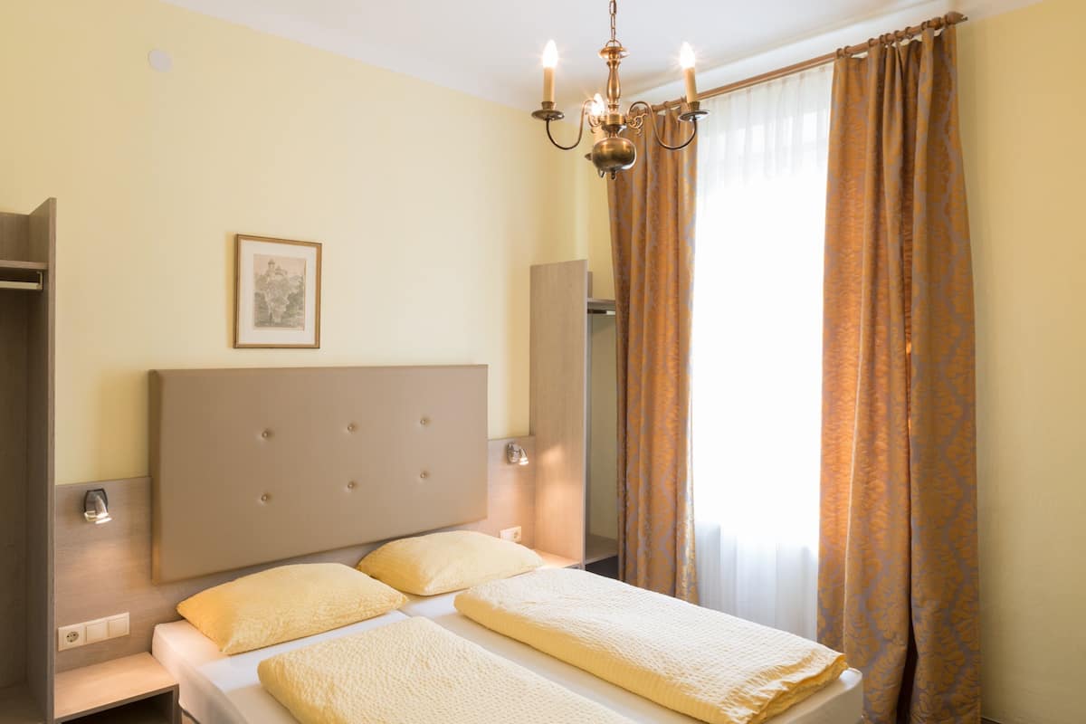 A hotel room with a large bed, a bedside table, a nightstand, a mirror, a chandelier, and a window with curtains.