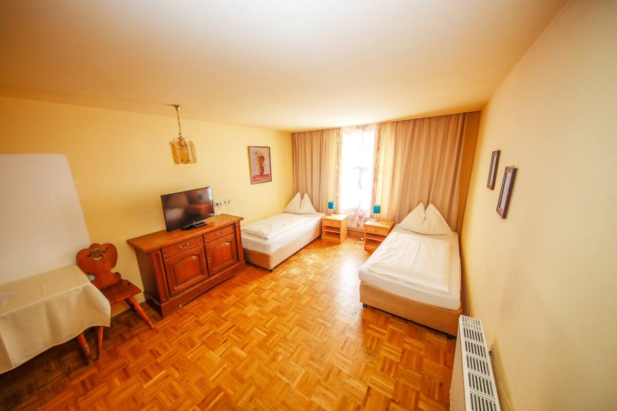A hotel room with a wooden parquet floor, a television, a desk with a chair, two single beds, a wardrobe, and a radiator.