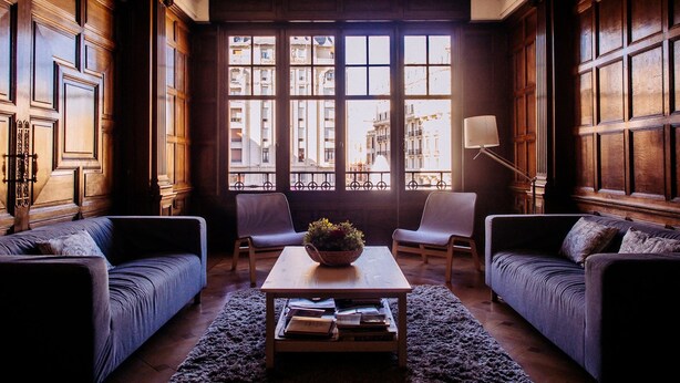 A room with two sofas, a coffee table with books, a lamp, and a view through large windows. - Zentric Hostel (León)