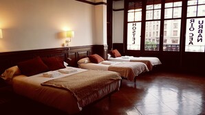A hotel room with two beds, a wooden headboard, a window with a view of buildings, and a wall-mounted lamp. - Zentric Hostel (León)