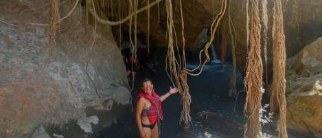 Una persona de pie en una cueva con agua, rodeada de grandes rocas y raíces colgantes.