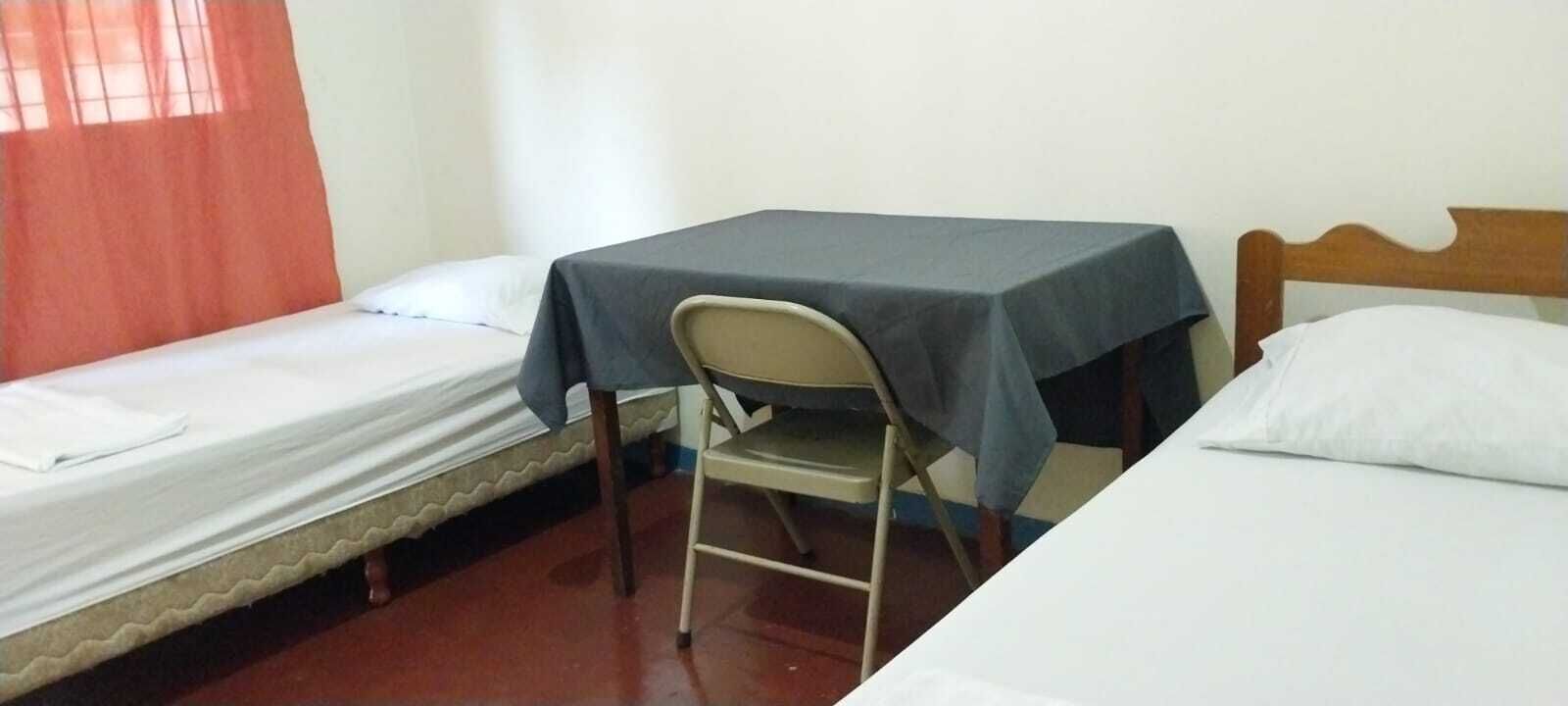 A room with two single beds, a small table with a tablecloth, a chair, and a window with curtains.