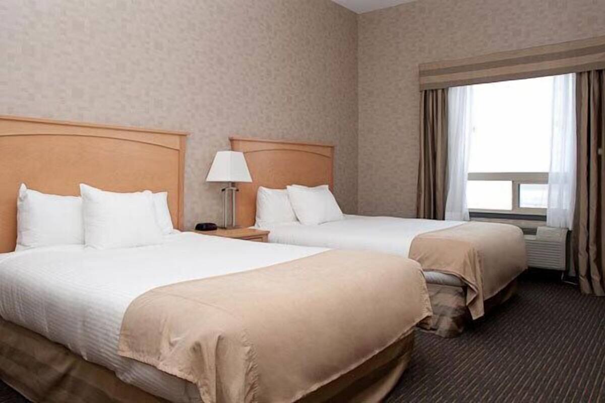 A hotel room with two beds, a nightstand with a lamp, and a large window with curtains.