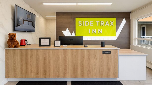 Una recepción de hotel con un letrero que dice 'SIDE TRAX INN', un peluche de oso, una taza roja, certificados enmarcados y un monitor LG.