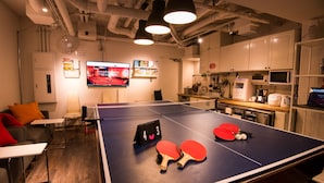 A ping pong table with two red paddles and a scoreboard in a room with a TV, kitchenette, and seating area. - Senba Hostel (Osaka)