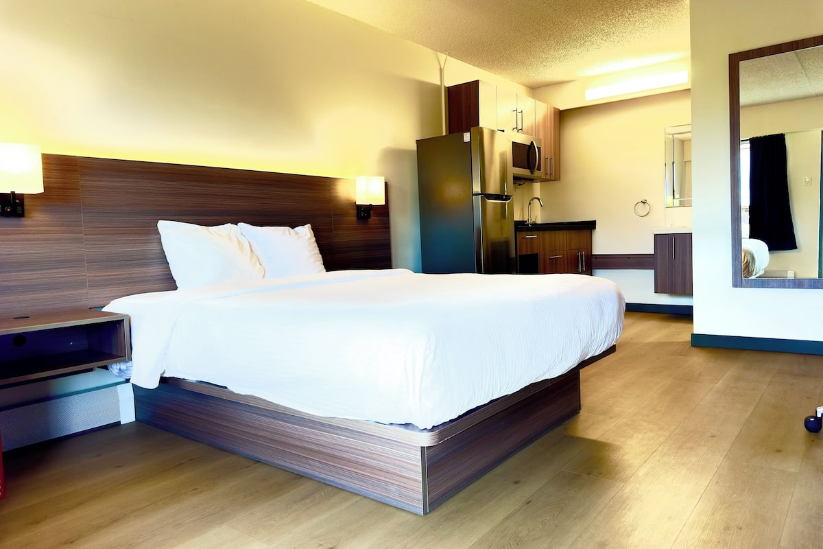 Een hotelkamer met een groot bed, een houten hoofdbord, een nachtkastje, een bureau, een spiegel, een kledingkast en een kitchenette.