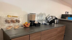 En köksbänk med en kaffemaskin, en fruktskål och en genomskinlig plastbehållare med bakverk.