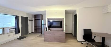 A modern hotel room with a wooden floor, a desk with a chair, a TV, a minibar, and a wardrobe.