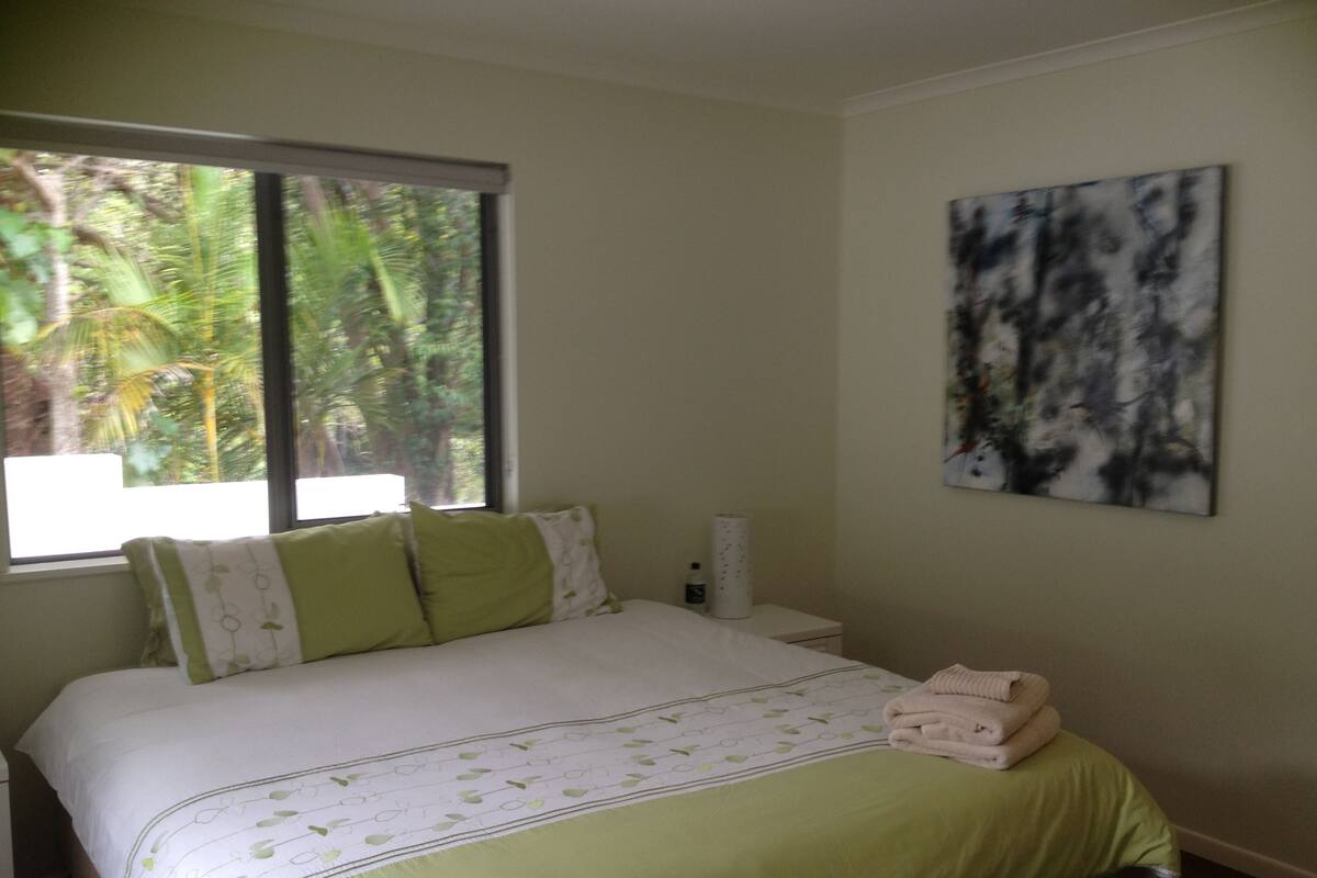 A bedroom with a bed, pillows, a bedside table, a lamp, and a window with a view of greenery.