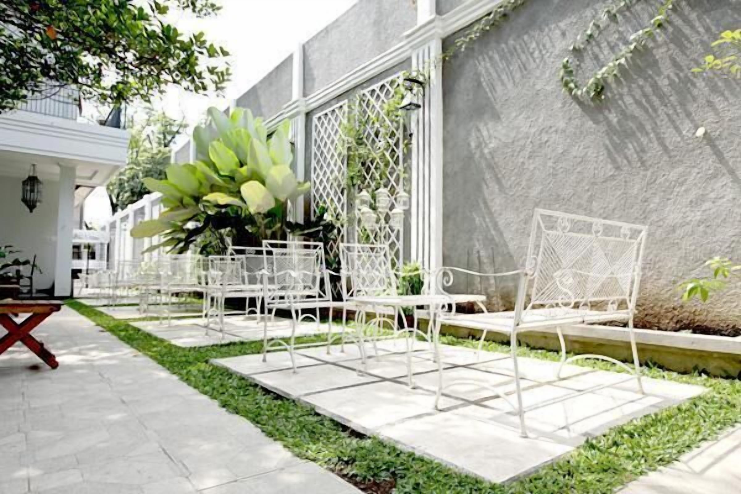 A patio area with white metal chairs and a dining table, surrounded by a small garden with a large plant and a white fence.