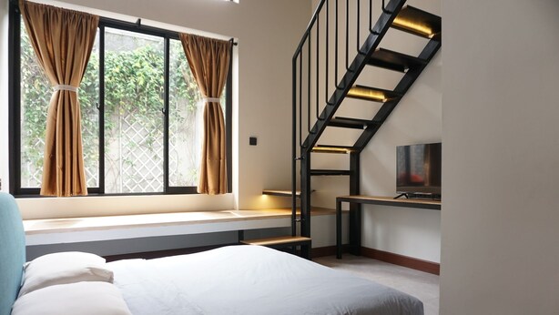 A bedroom with a bed, a window with curtains, a staircase, and a TV on a stand. - De Halimun Guest House (Bandung)