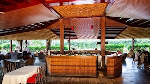 Un restaurante al aire libre con mesas y sillas de madera, una zona de bar central y vistas a zonas verdes.