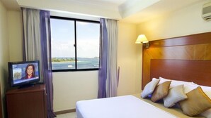 A hotel room with a bed, a television on a wooden stand, and a view of the sea through a large window. - Mookai Hotel (Male)