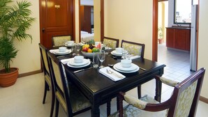 A dining area with a long table set for six, surrounded by chairs. A potted plant is on the left, and a view of the kitchen is visible through an open door. - Mookai Hotel (Male)