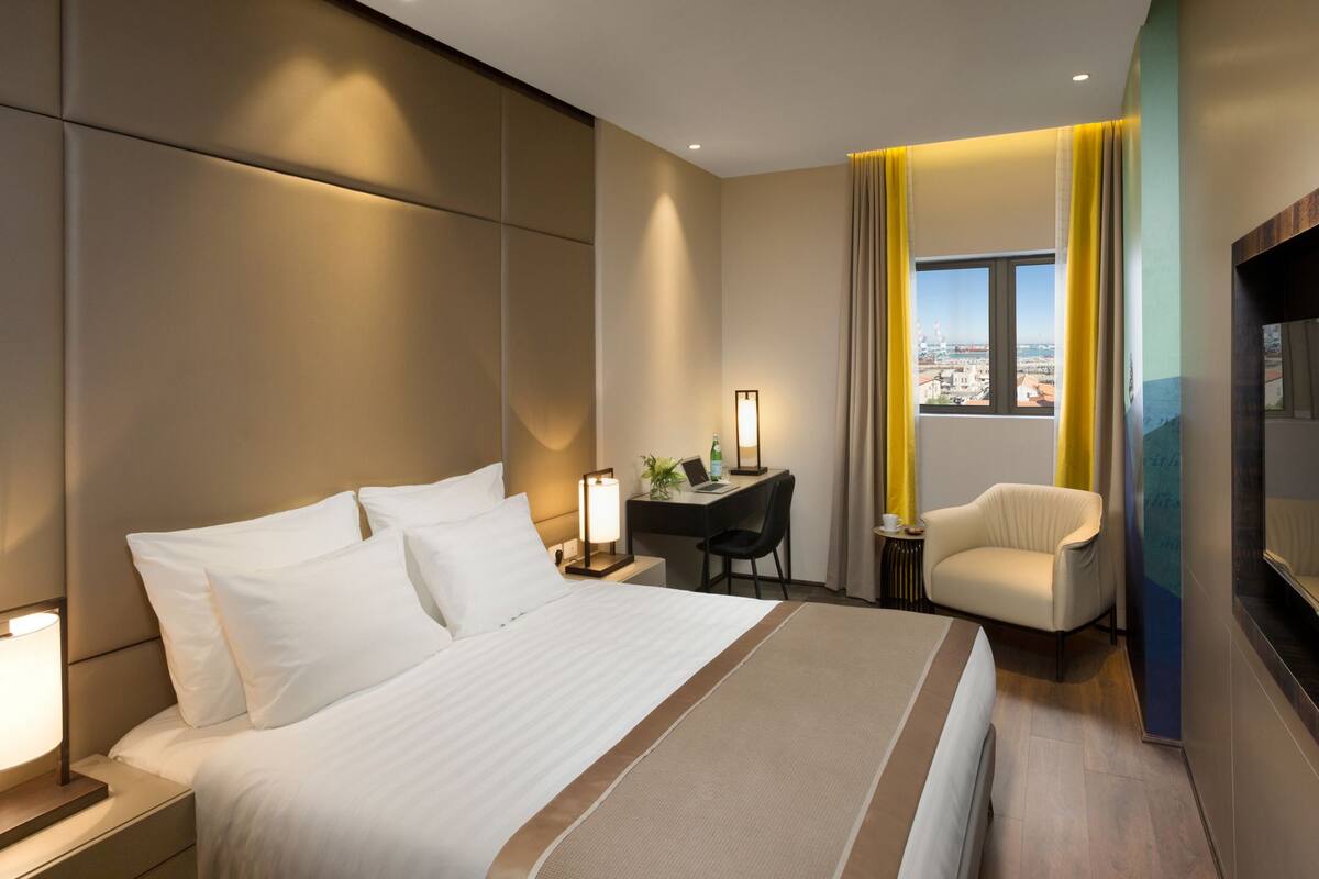 Una habitación de hotel moderna con una cama grande, un escritorio con silla, un televisor y vistas a la ciudad a través de una ventana.