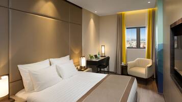 Una habitación de hotel moderna con una cama grande, un escritorio con silla, un televisor y vistas a la ciudad a través de una ventana.