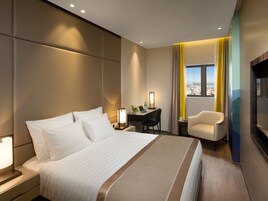 Una habitación de hotel moderna con una cama grande, un escritorio con silla, un televisor y vistas a la ciudad a través de una ventana.