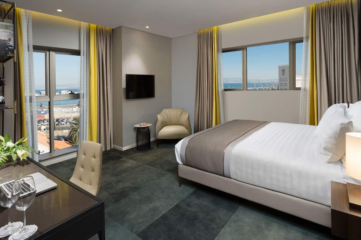 Una habitación de hotel con una cama, un escritorio con una computadora portátil y gafas, una silla, una mesita y un televisor en la pared.