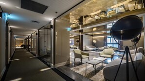 Eine moderne Hotelhalle mit Glastrennwand, einer Sitzecke mit Sofas und Sesseln sowie einer Decke mit freiliegenden Lüftungsrohren.