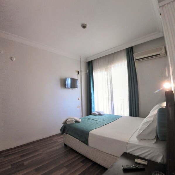 A hotel room with a bed, a TV mounted on the wall, an air conditioning unit, and a window with curtains.