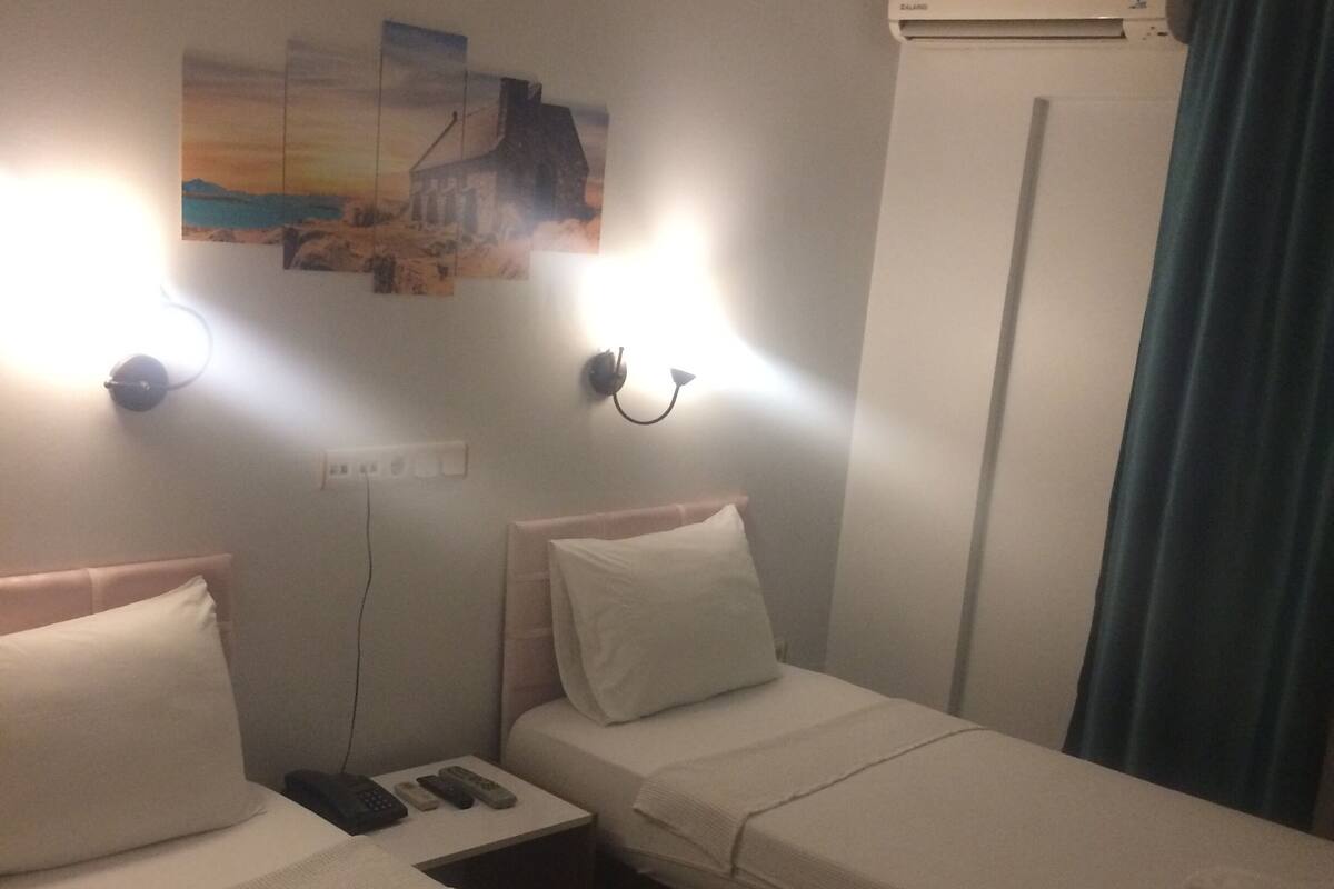 A hotel room with two single beds, a nightstand with a phone and remote controls, a wall-mounted air conditioner, and a painting above the beds.