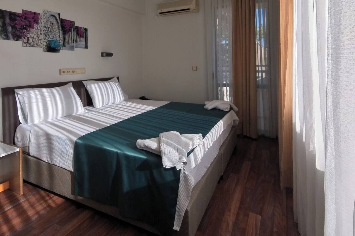 A hotel room with a bed, a bedside table, an air conditioning unit, and a window with curtains.