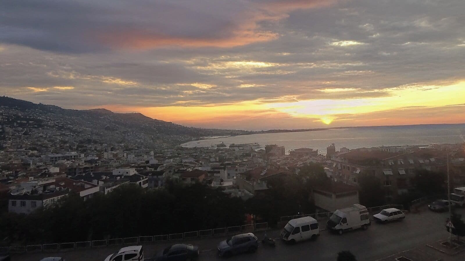A coastal cityscape at sunset with buildings, a road, and parked vehicles.