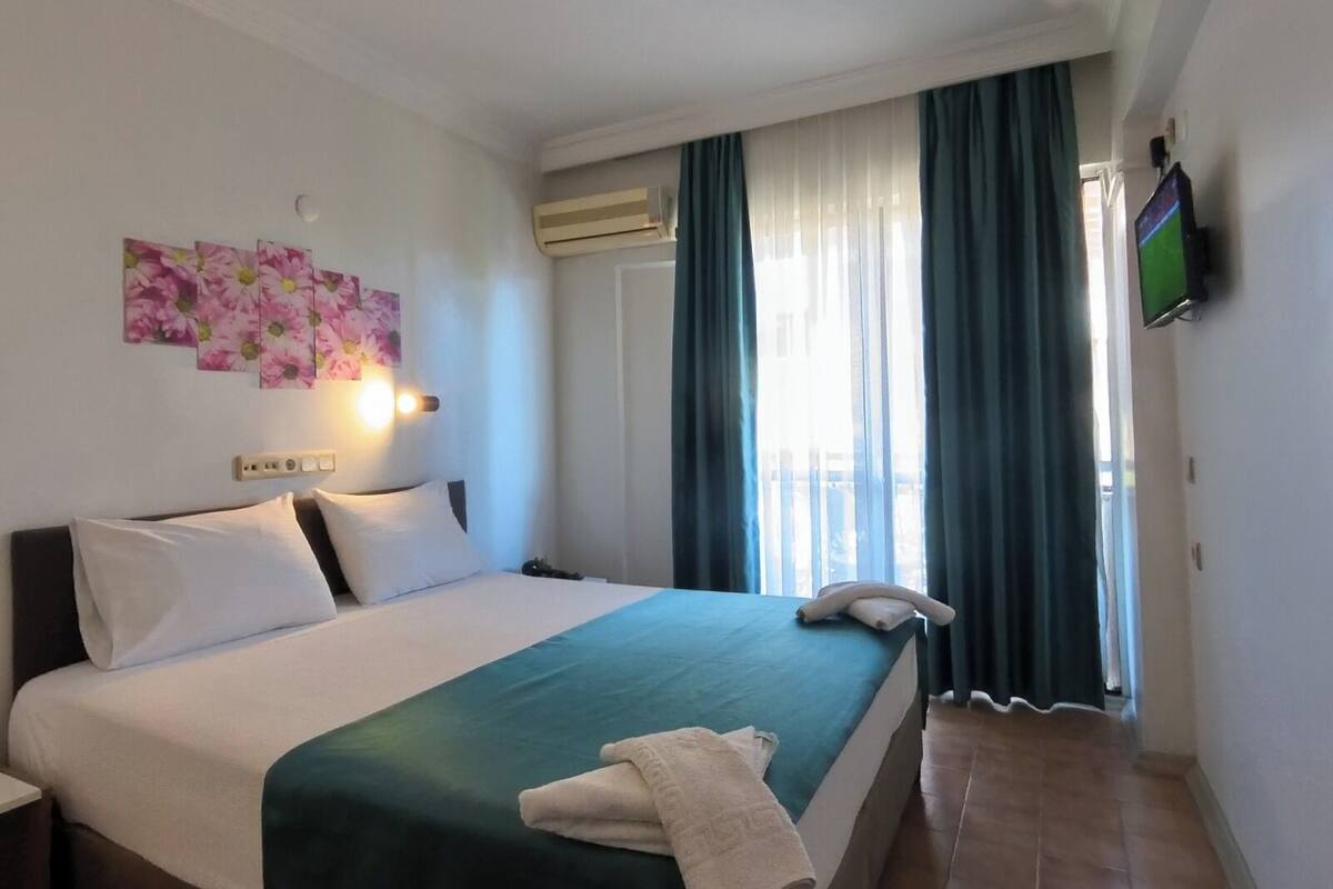A hotel room with a bed, a flat-screen TV, an air conditioning unit, and a window with curtains.