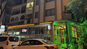 A multi-story building with a ground-level commercial space featuring a glass facade and illuminated signage. - HBS Hotel (Kusadasi)
