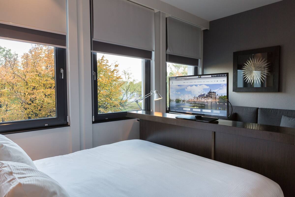 A modern hotel room with a large bed, a desk with a computer, and a view of autumn trees through the windows.