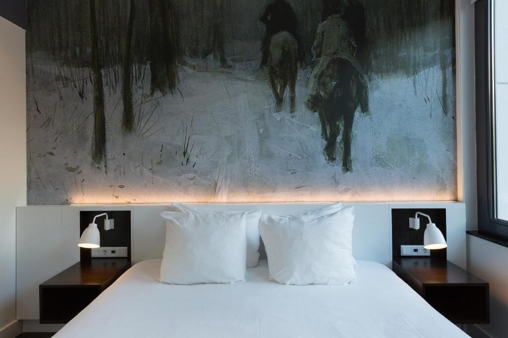 A modern hotel room with a large abstract painting on the wall, a neatly made bed with white linens, two bedside tables with lamps, and a window providing natural light.