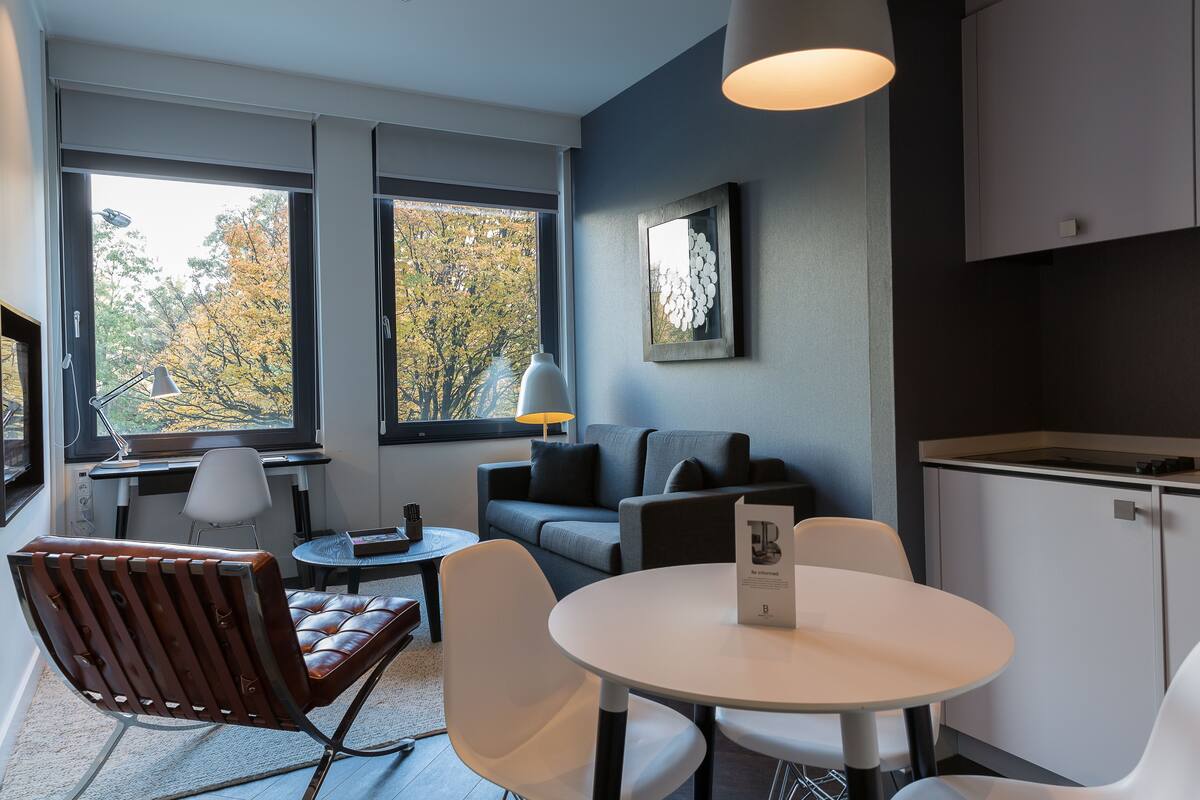 A modern room with a sofa, chairs, a table, and a view of autumn trees through large windows.