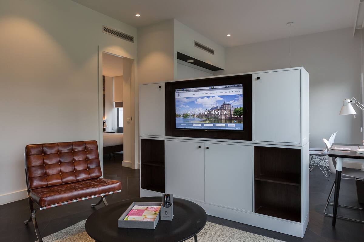 A modern hotel room with a large flat-screen TV mounted on a white cabinet, a brown leather sofa, a small table with a magazine, and a desk with a lamp.